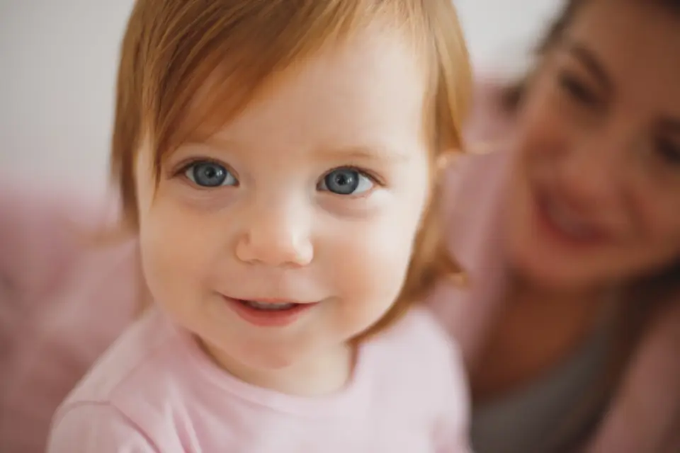 A small child smiling at the camera, a parent softly out of focus behind